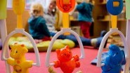 Kinderspielzeug hängt an einem Rahmen. Im Hintergrund spielt eine Erzieherin mit zwei kleinen Kindern (dpa / Julian Stratenschulte)